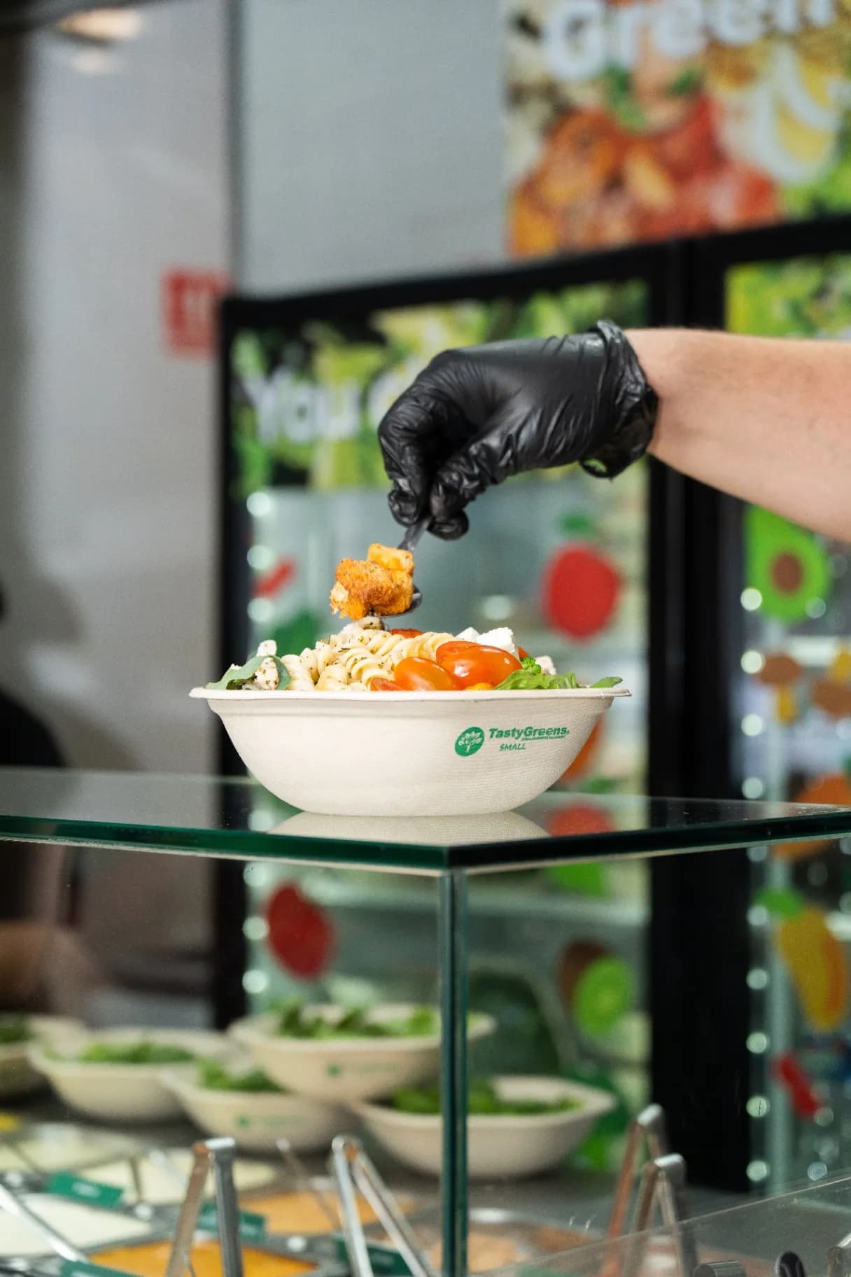 Mano con guante negro dando el toque final a un bowl Tasty Greens detrás de la barra